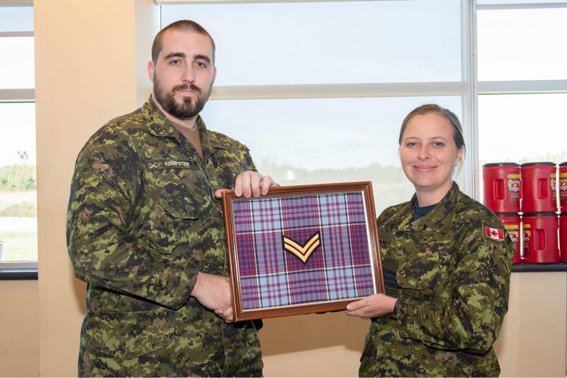 Kempster in Canadian CADPAT uniform holding a framed corporal chevron beside a fellow service member, October 2019.